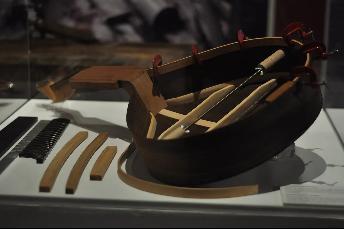 An unfinished Portuguese guitar body in a museum case, with its ribs clamped and a maker's tools beside it.